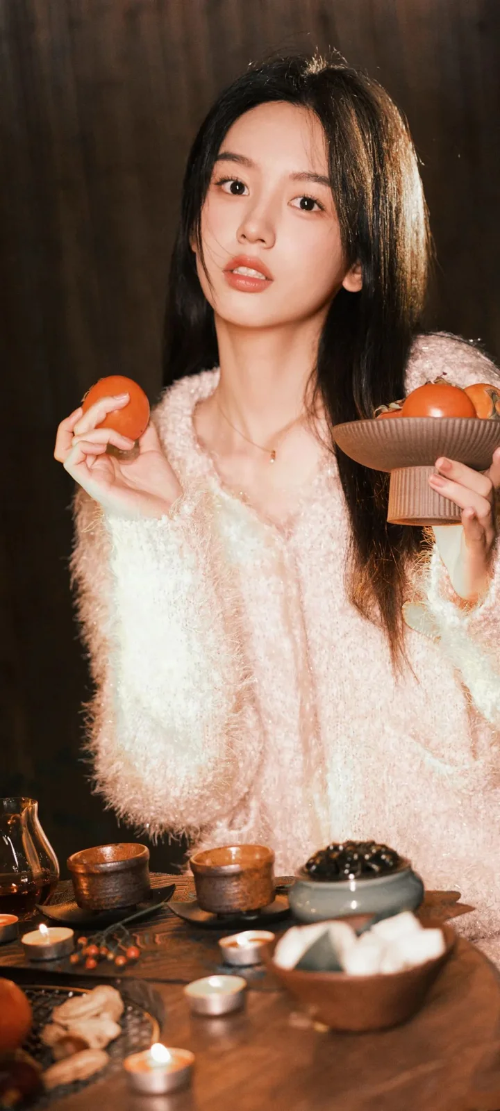긴 머리의 여성이 밝은 색 스웨터를 입고 따뜻한 조명 아래에서 오렌지와 과일 접시를 들고 차 테이블 앞에 앉아 있습니다.