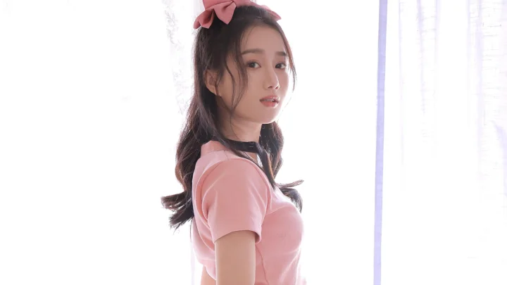 一位戴着粉色蝴蝶结、穿粉色上衣的成年女性站在明亮窗边。