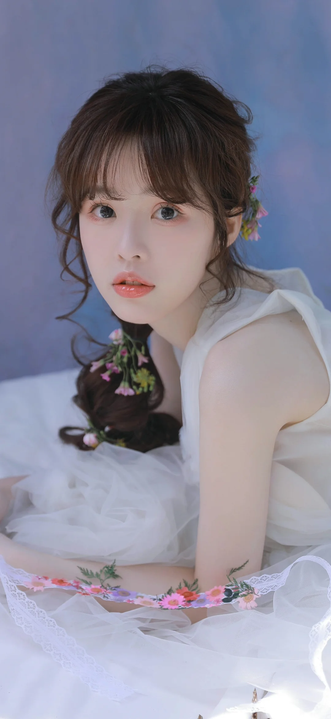 一位身穿白色连衣裙、佩戴花饰的女性坐在浅蓝色背景前。