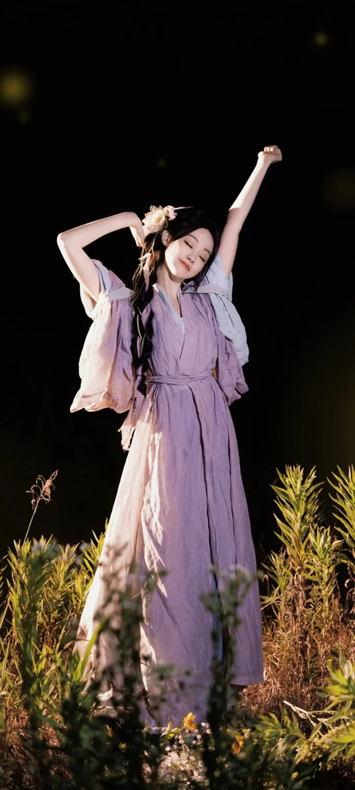 一名身穿淡紫色连衣裙的女子站在夜色中的植物与野花之间。
