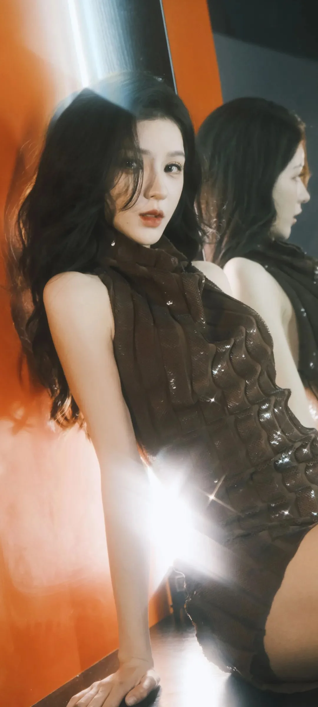어두운 드레스를 입은 성인 여성이 거울 앞에 서 있으며 정면과 옆모습의 반사가 함께 보입니다.