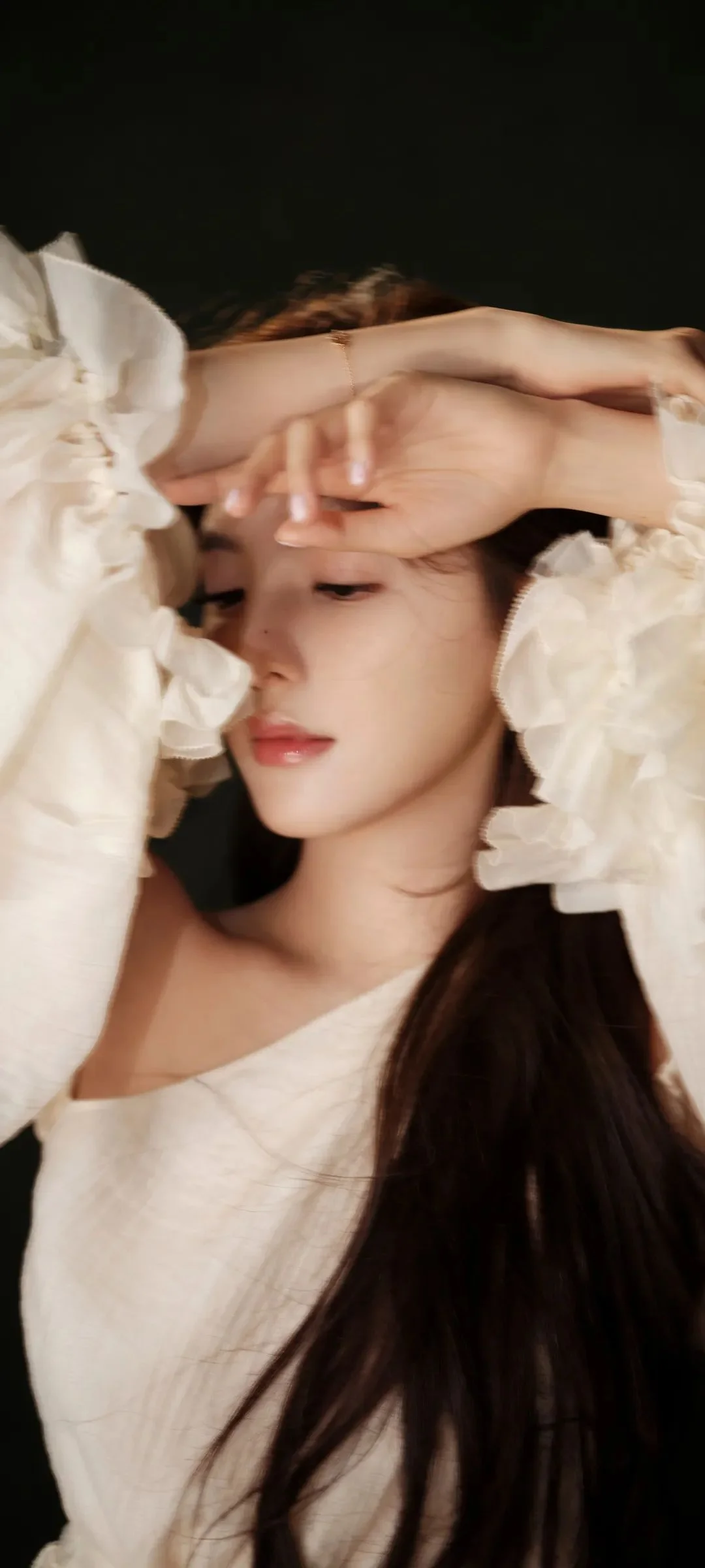 긴 머리의 여성이 흰색 드레스를 입고 어두운 배경 앞에서 팔을 이마에 올리고 있다.