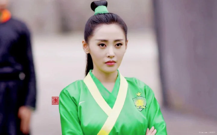 一名盘发女子身穿绿色服饰站在建筑背景前。