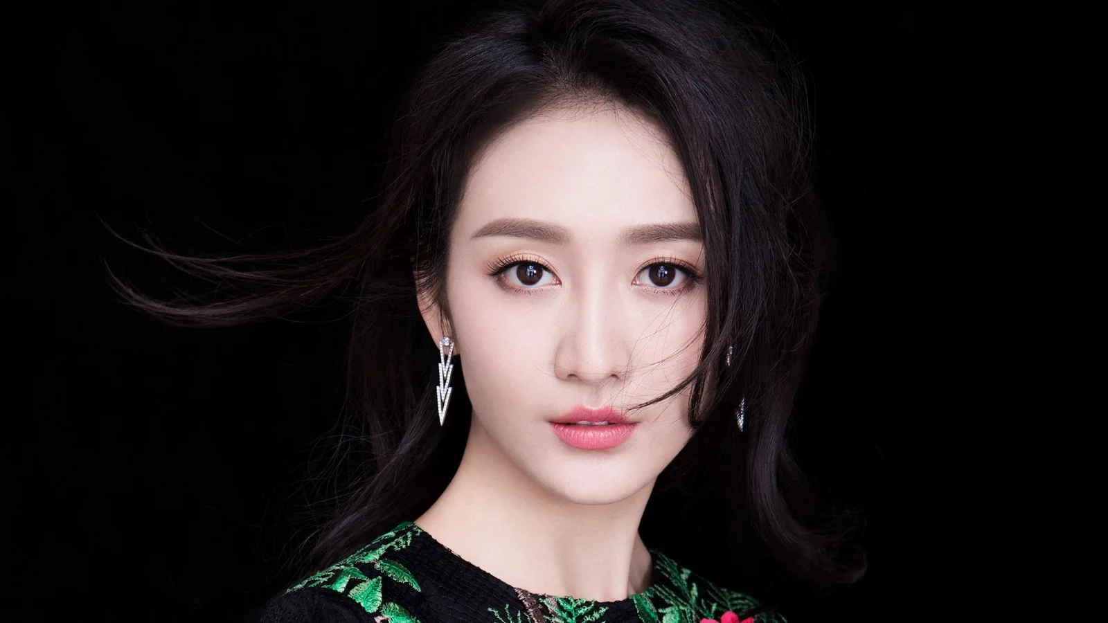 黑色背景前，一位深色长发女性直视镜头，佩戴银色耳饰并穿着带绿色图案的黑色上衣。
