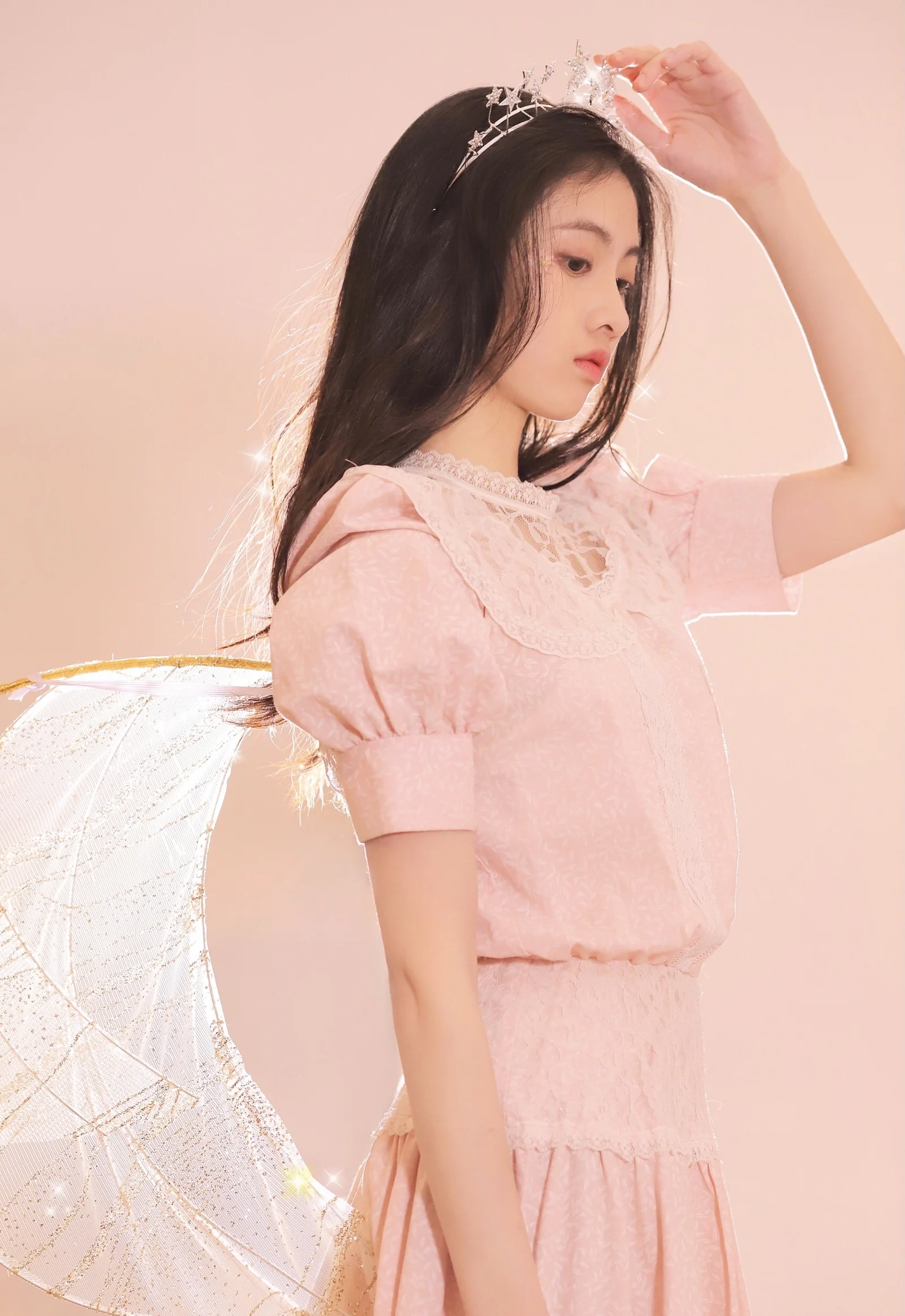 ピンクのドレスときらめく髪飾りを身につけ、翼のような装飾を背負った女性がピンクの背景の前に立っています。