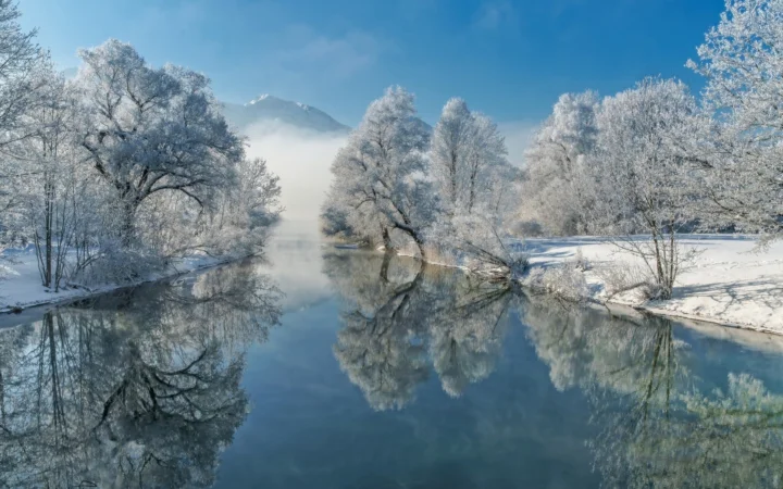 覆满白霜的树木环绕着宁静河流，远处山峦隐现在薄雾中，水面映出冬日景色。