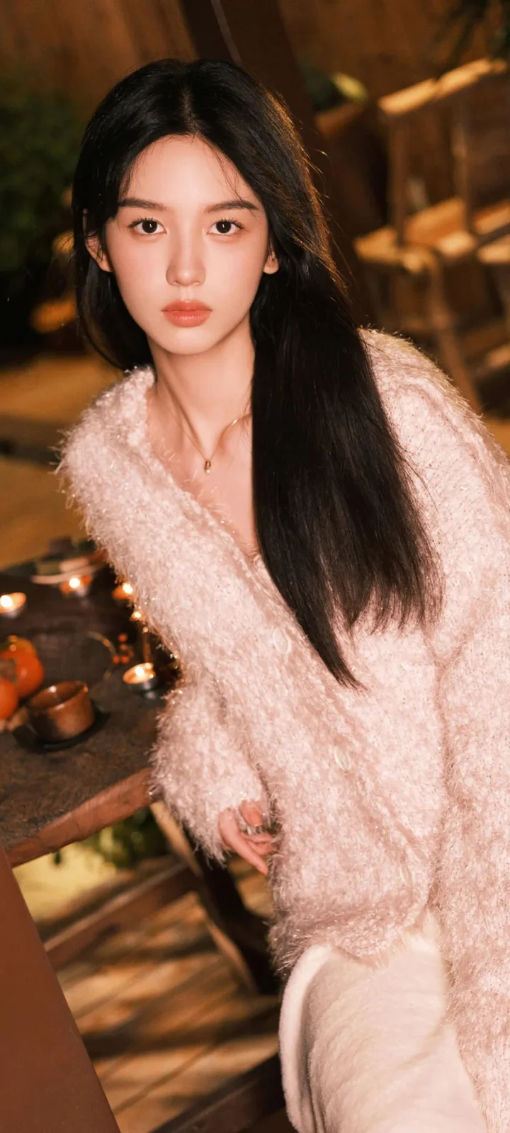 一位長髮女性穿著粉色毛衣站在溫暖燈光下的室內環境中。