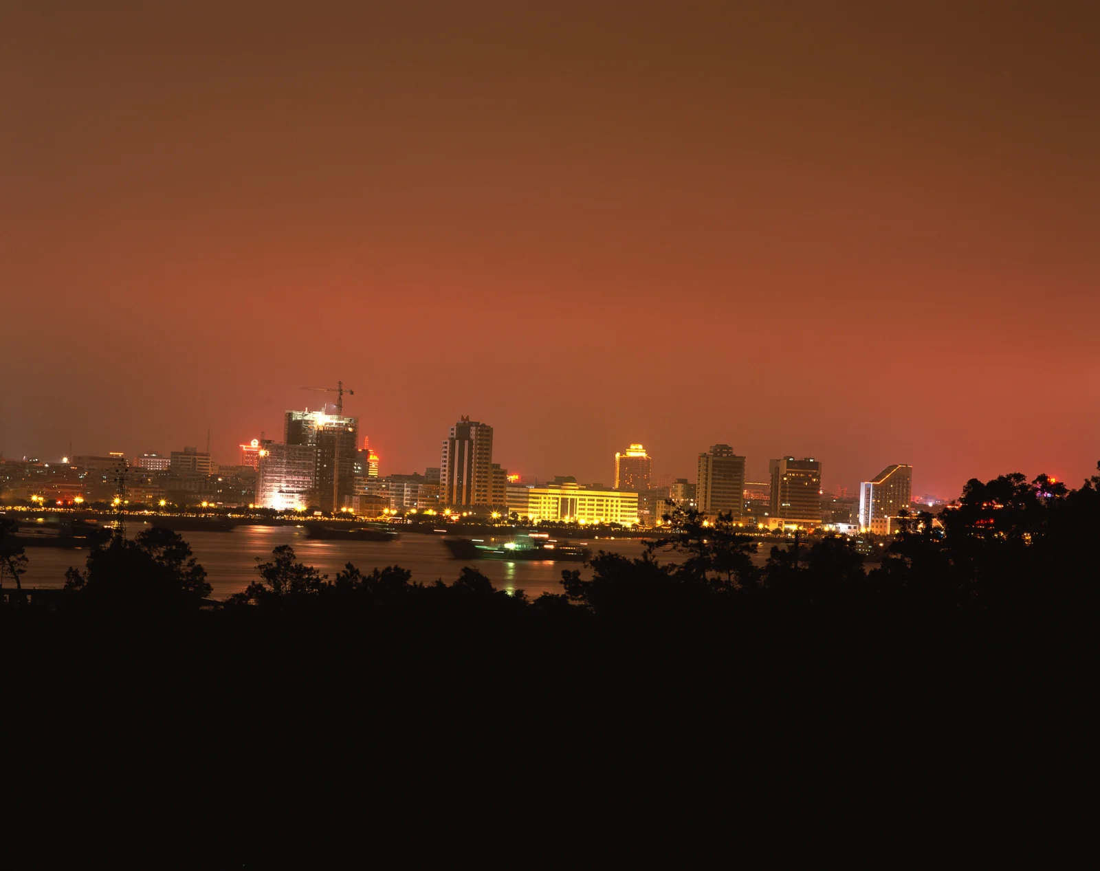 夜色中的城市建筑沿水岸延伸，前景是树木剪影，天空呈现深橙色。