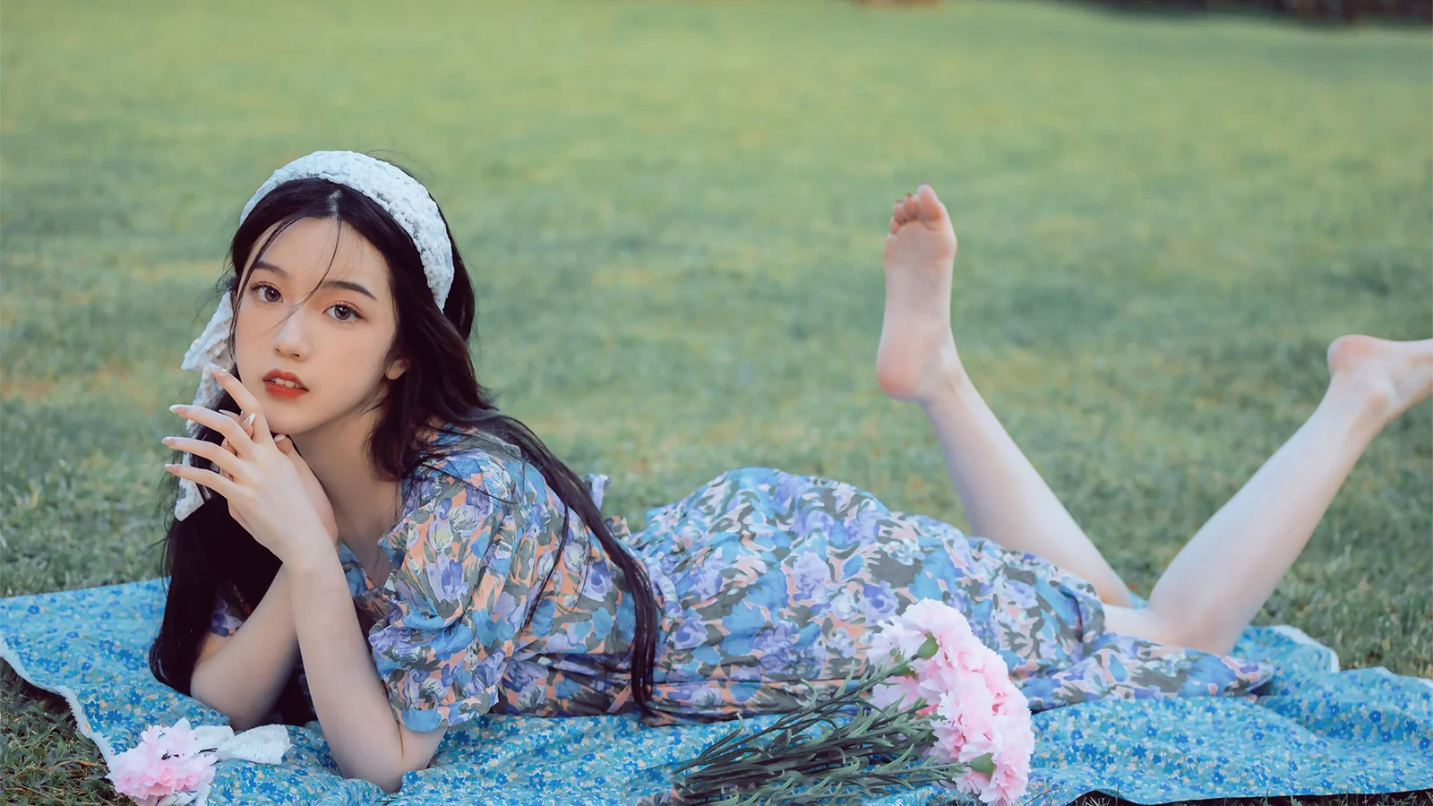 花柄のワンピースを着た女性が、ピンクの花のそばで青いブランケットの上に横たわっています。