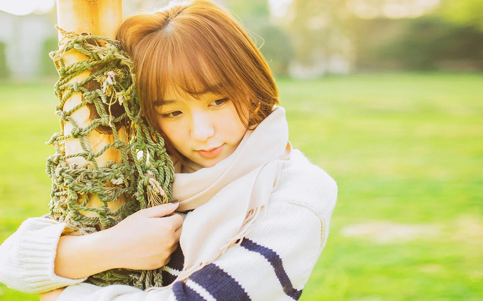 흰 목도리를 두른 여성이 푸른 잔디 옆에서 짜인 그네를 안고 있다.