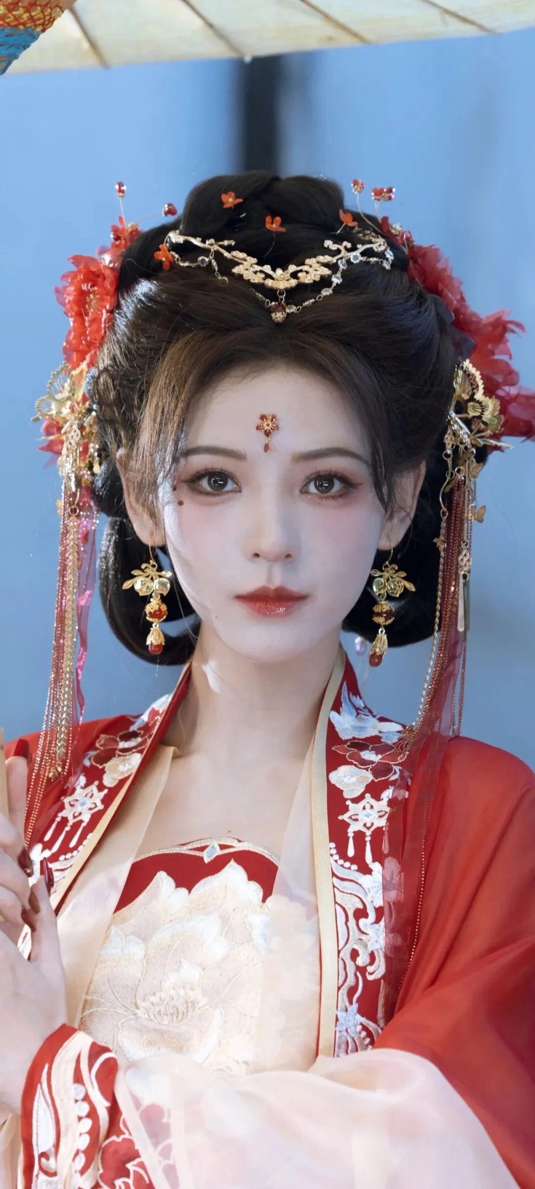 青い背景の前で、華やかな髪飾りを着けた赤と白の伝統衣装の成人女性がカメラに向かっている。