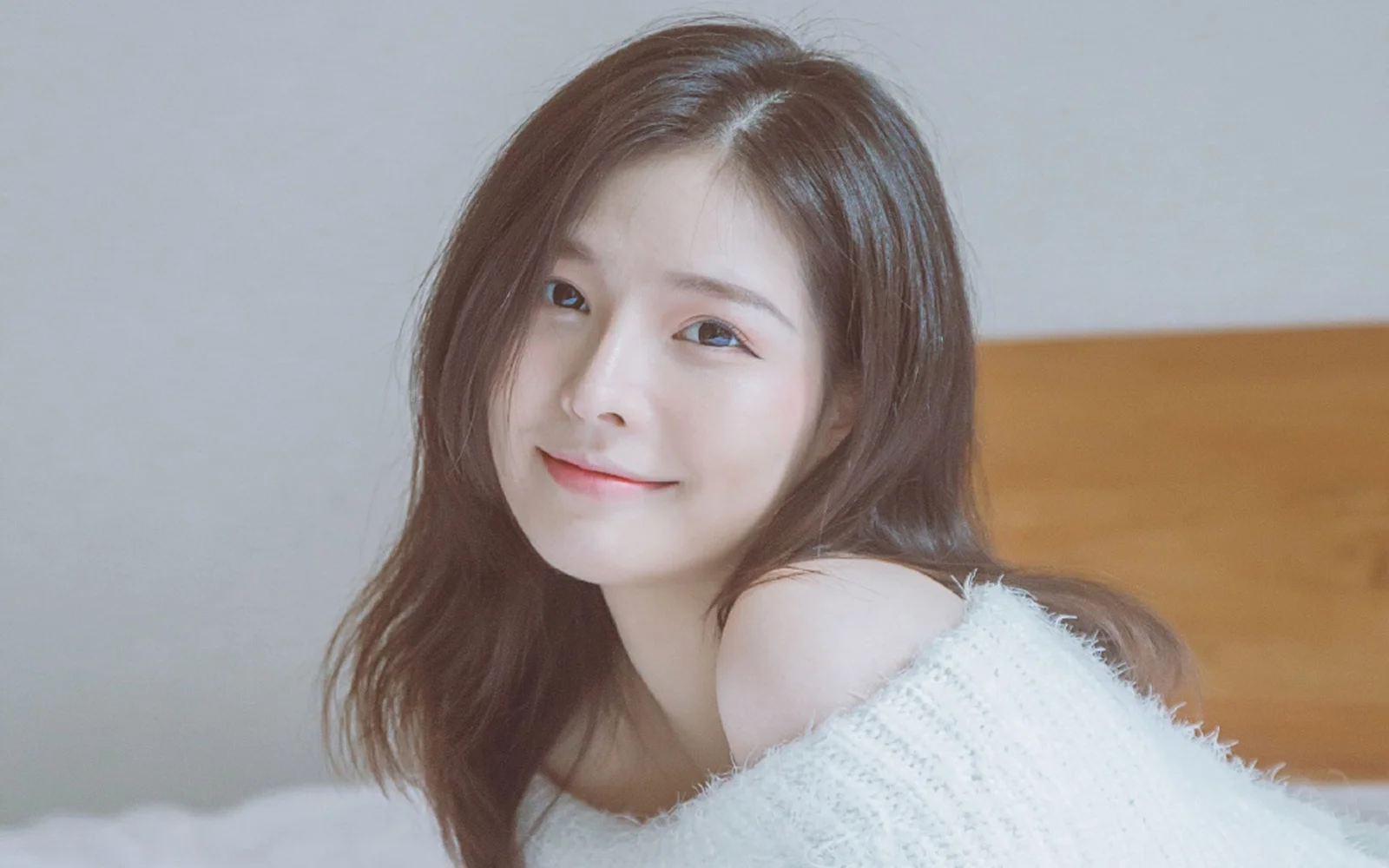 긴 갈색 머리의 여성이 카메라를 향해 미소 짓고 있으며 뒤로 밝은 벽과 나무 배경이 보입니다.