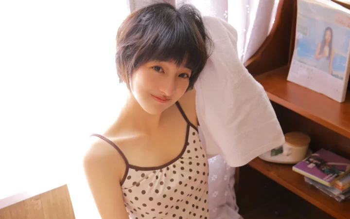一名短发女性在室内靠近明亮窗户的位置微笑，头上搭着一条白色毛巾。