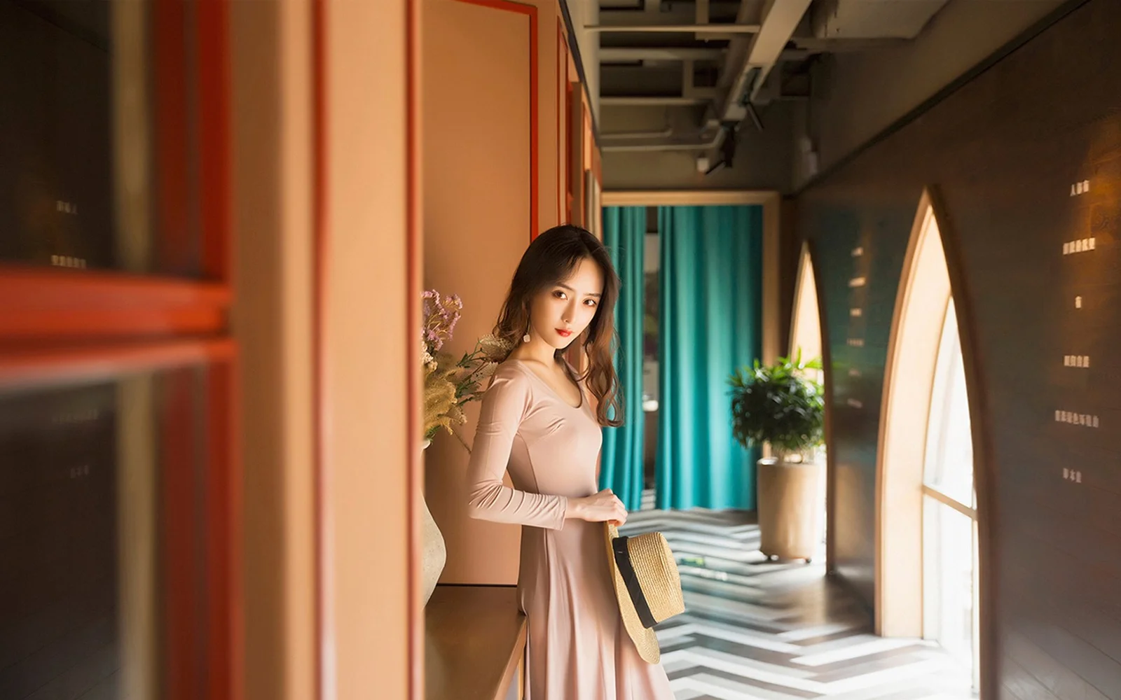 一位身穿浅色长裙的成年女性站在带有拱形窗和蓝绿色窗帘的室内走廊中。
