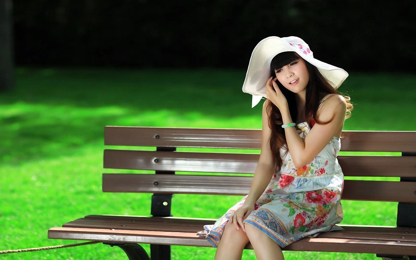 つばの広い帽子をかぶった女性が、緑の芝生のそばの公園のベンチに座っています。