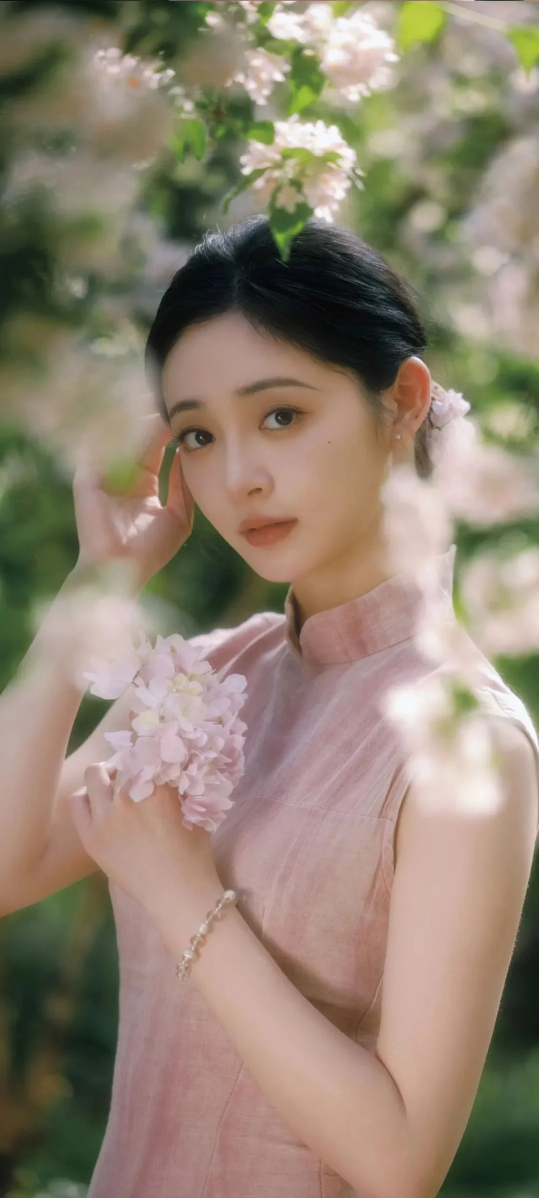 분홍색 옷을 입은 여성이 꽃이 핀 나뭇가지와 부드러운 녹음 속에서 연한 색의 꽃다발을 들고 있습니다.