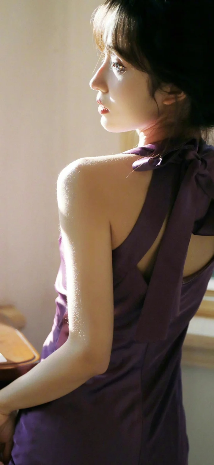 一名成年女性穿着紫色连衣裙侧身回望，室内柔和光线勾勒出她的轮廓。