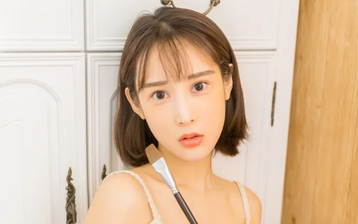 一位短发女性手持化妆刷站在室内白色柜门前。
