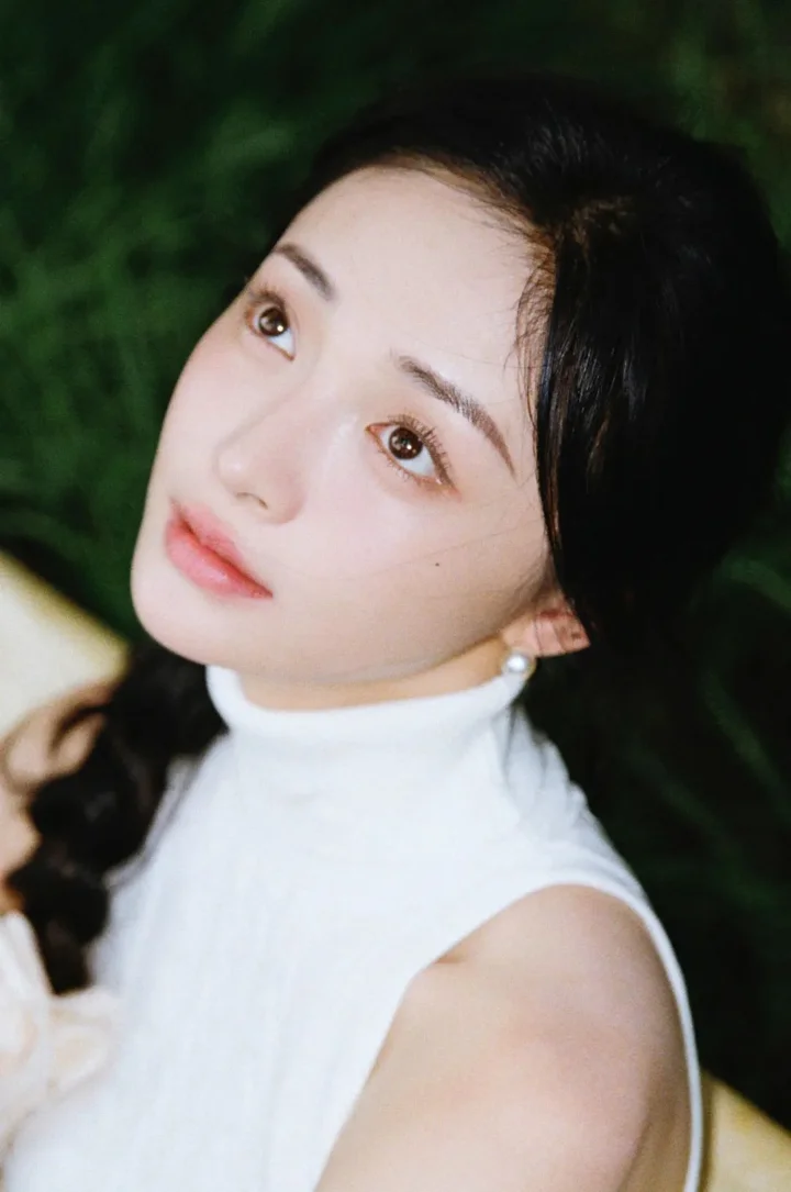 一名成年女性身穿白色无袖高领上衣，在深绿色植物背景前侧头望向镜头。