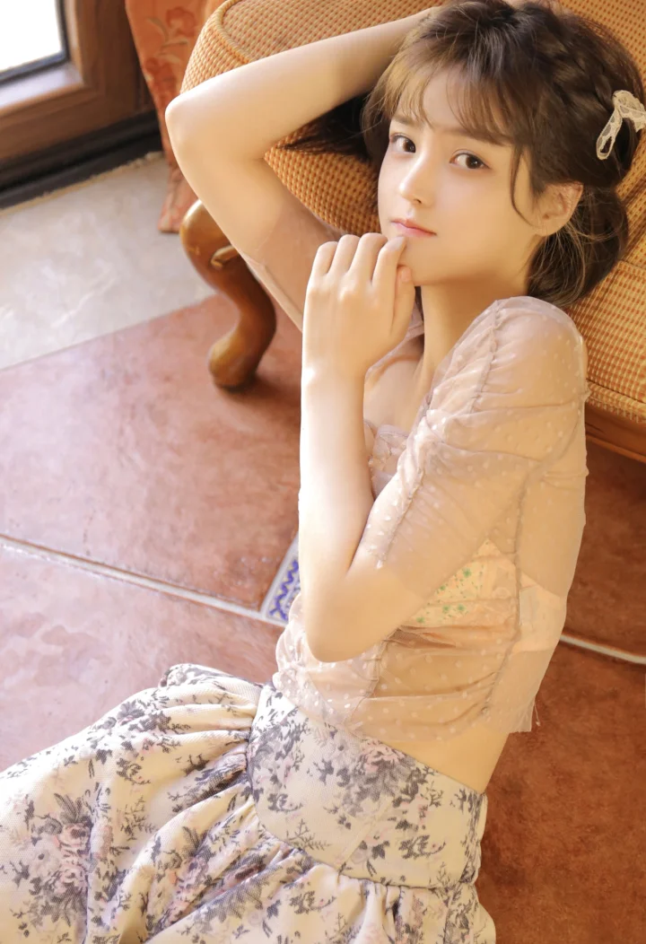 一位成年女性坐在椅子上，身穿浅色上衣和花卉长裙，面向镜头。
