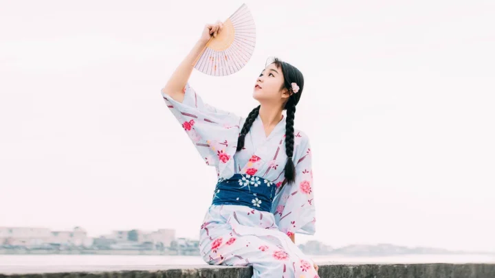 一名身穿浅色花卉和服、手持折扇的女子坐在水边。