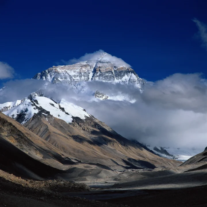 巍峨的积雪山峰穿过云层，前景是荒凉的山谷与冰川地貌。