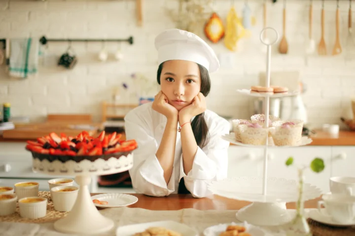 一名戴着白色厨师帽、穿着白色厨师服的女子在摆放着糕点的面包房柜台前托腮。