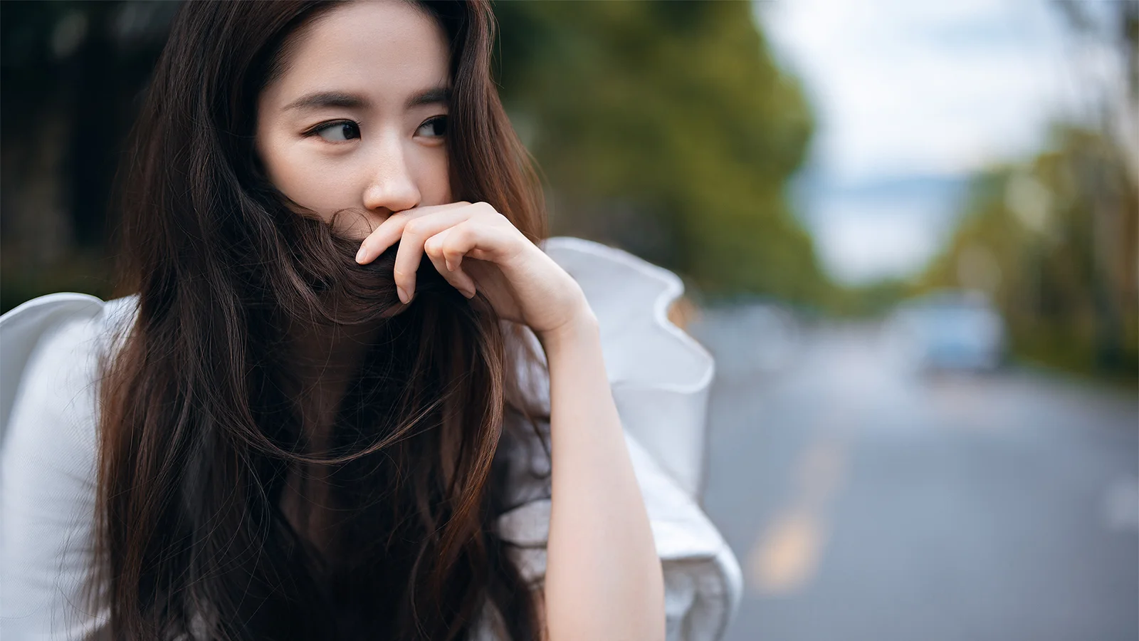 긴 머리의 여성이 야외 도로 옆에서 한쪽을 바라보고 있으며 배경에는 나무와 도로의 부드러운 흐림 효과가 보입니다.