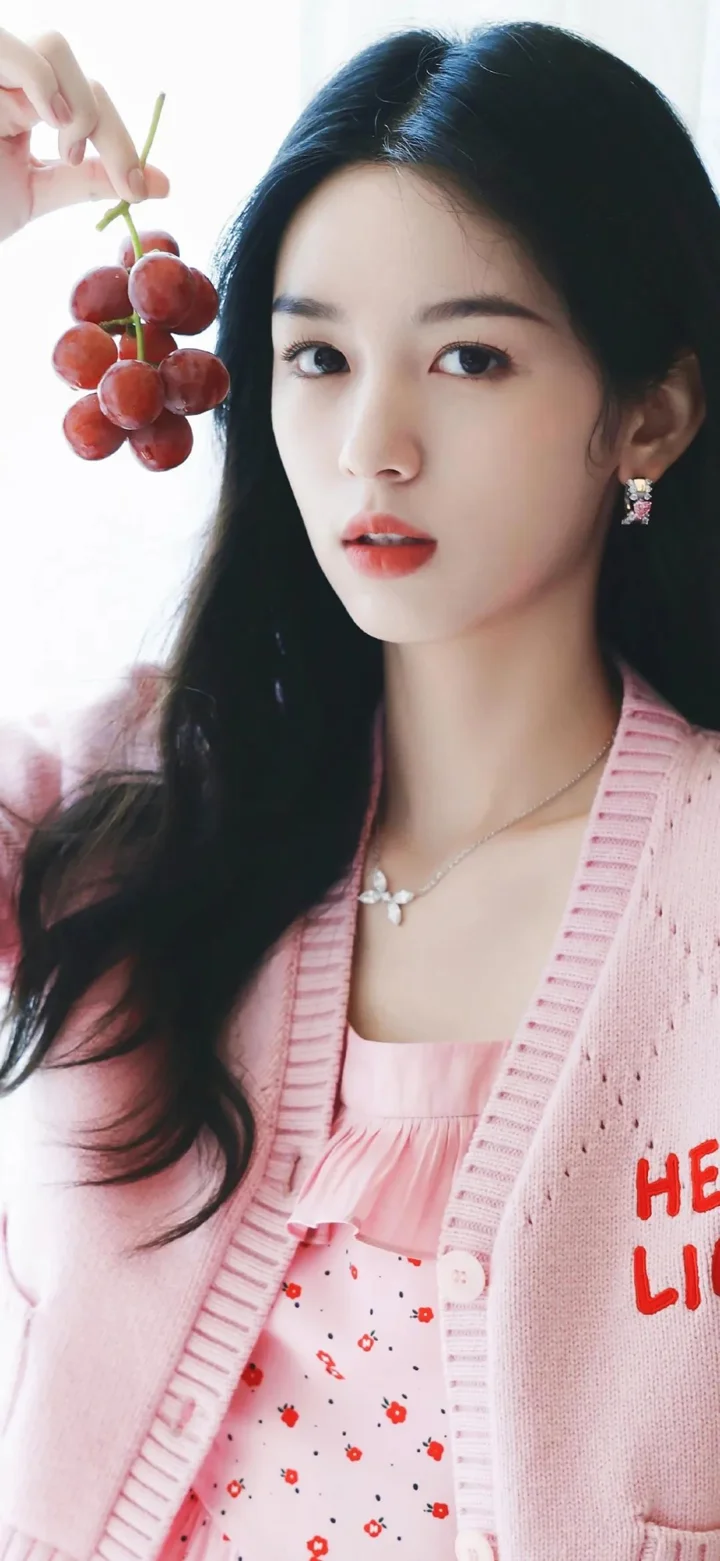 긴 머리 여성이 분홍색 카디건을 입고 포도 한 송이를 든 채 카메라를 바라보고 있습니다.
