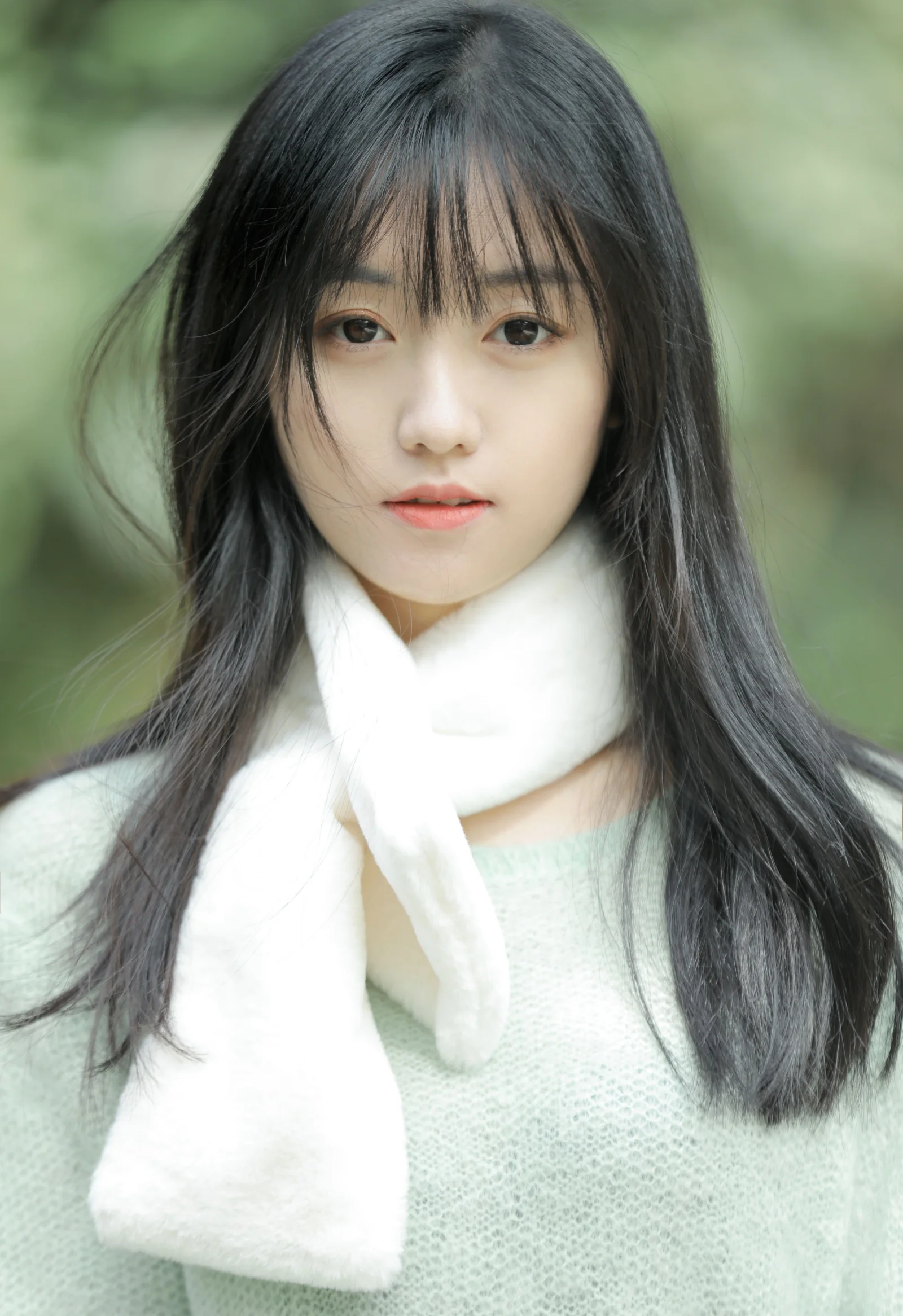 一名长发女子戴着白色围巾，站在柔和的绿色户外背景前。