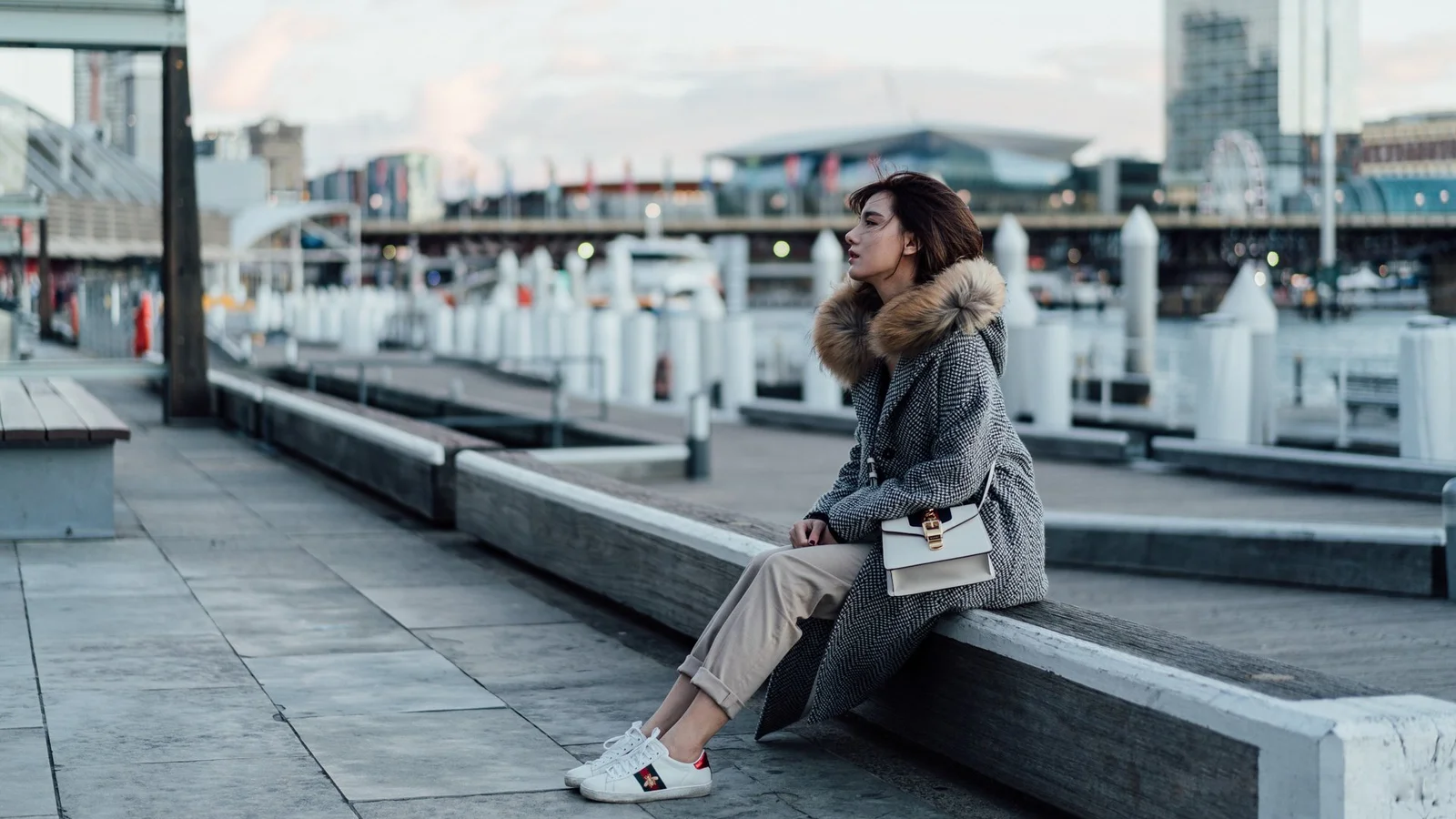 一名女子穿著灰色外套坐在濱水平台邊緣，身後是碼頭和城市建築。