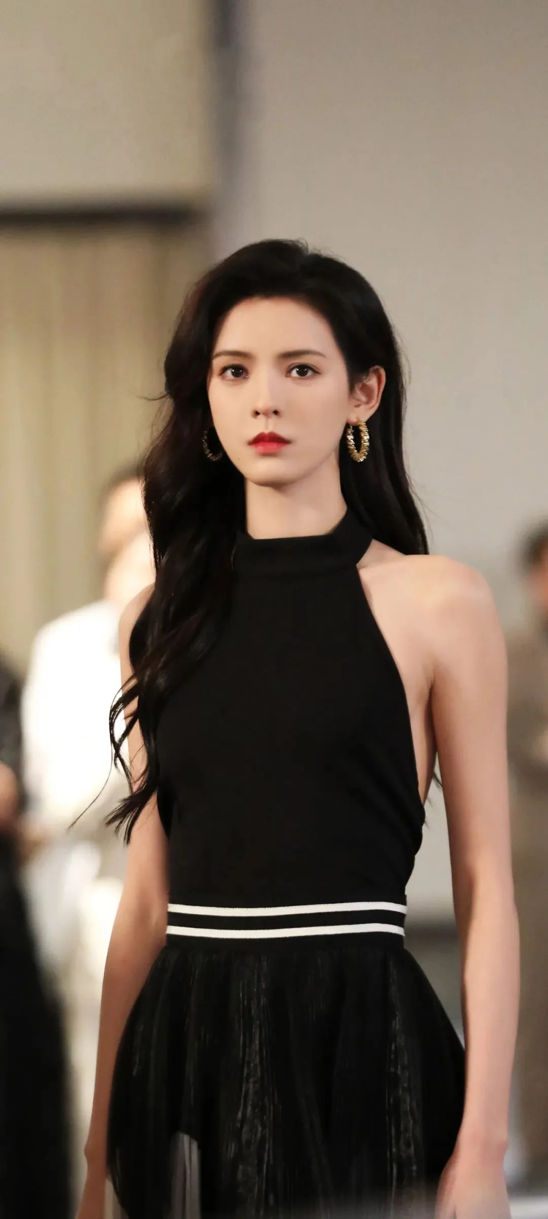 一名长发女性身穿黑色无袖连衣裙站在室内，背景呈现柔和的虚化效果。