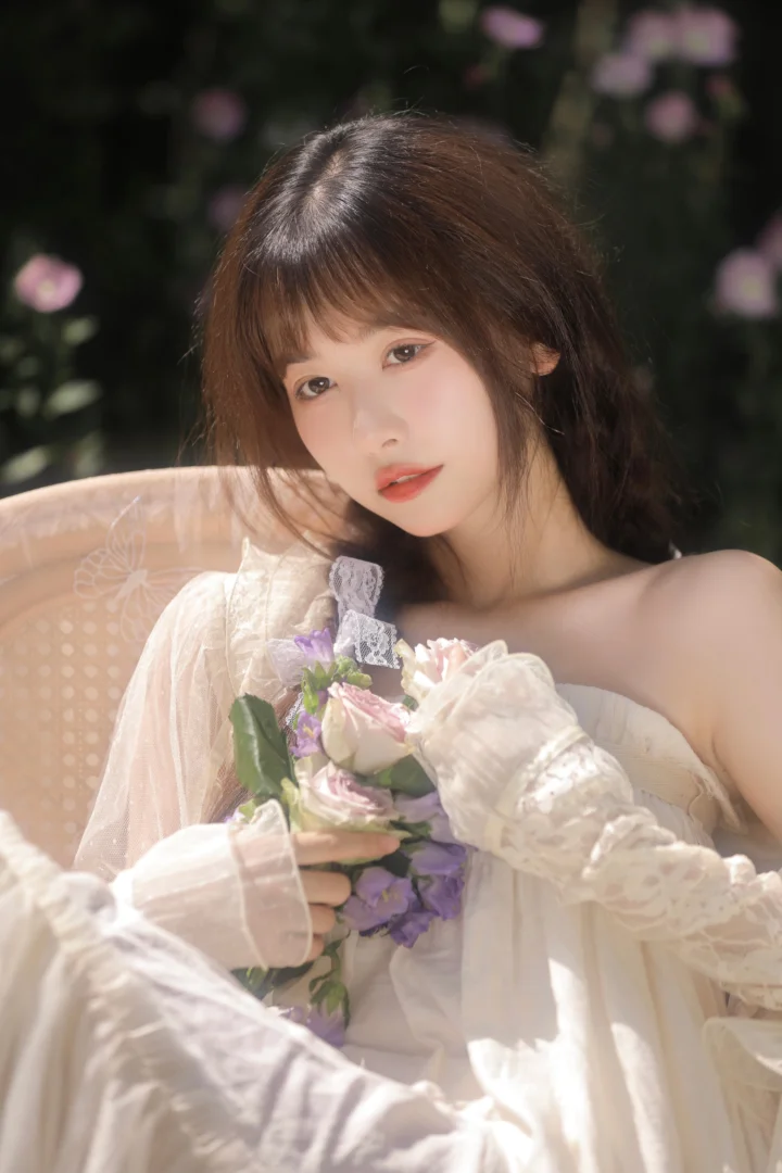 一位成年女性身穿白色蕾丝服装，手捧花束坐在椅子上，背景是虚化的花卉与绿叶。