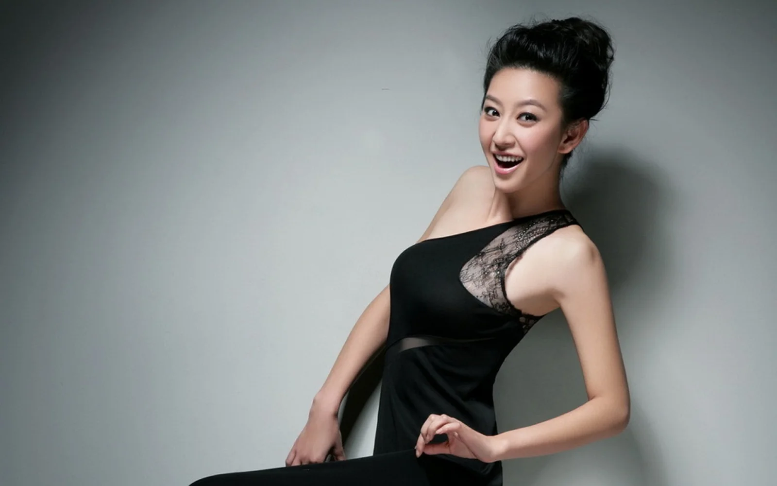 黒いドレスを着た成人女性が、グレーのスタジオ背景の前でポーズを取り微笑んでいる。
