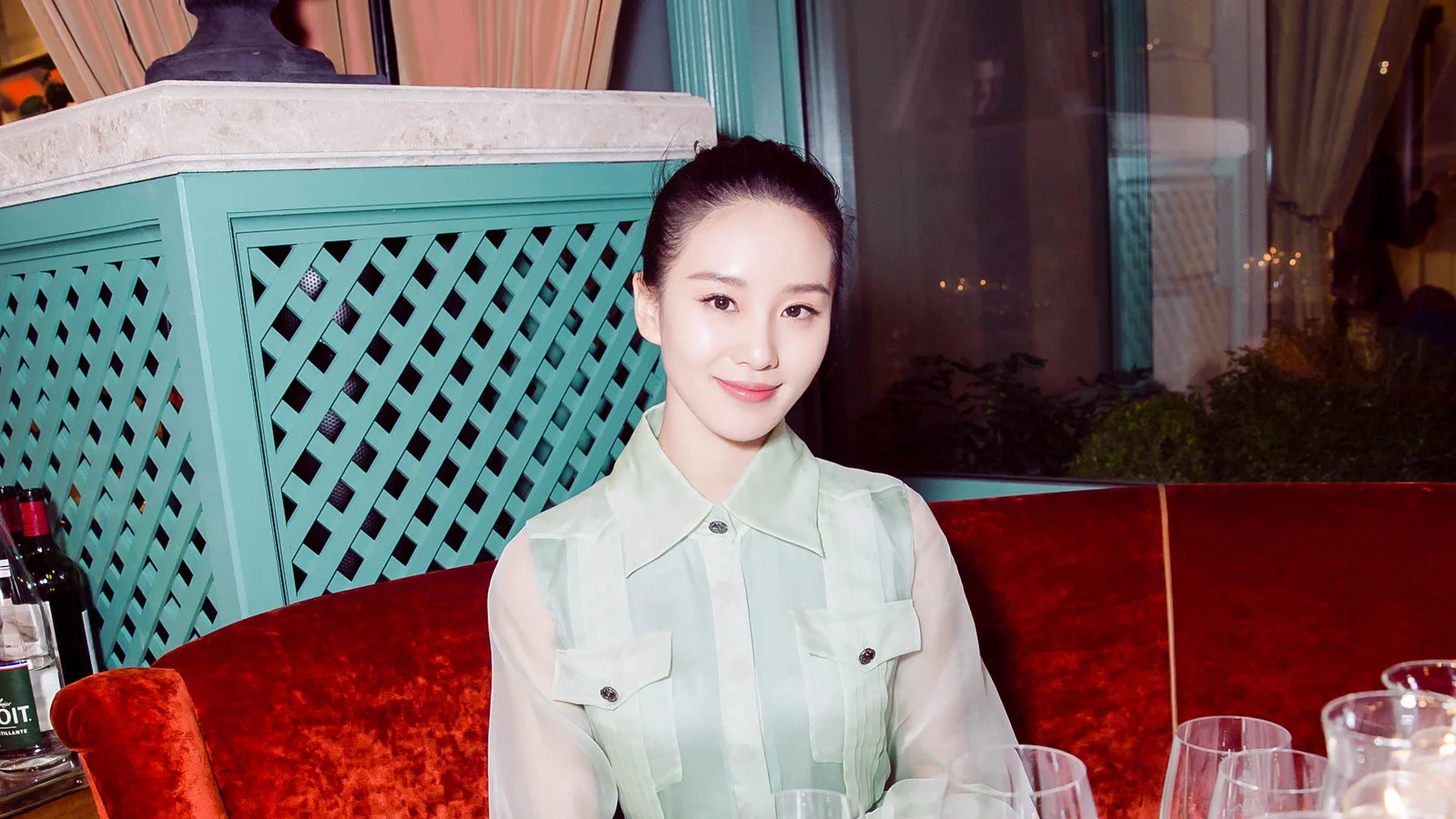 一名成年女性坐在餐厅红色沙发前，身后是青绿色格栅和玻璃窗。