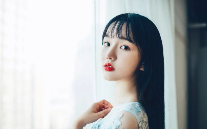검은 머리의 여성이 창가에서 카메라를 바라보고 있으며 부드러운 빛 속에서 밝은색 상의를 입고 있습니다.