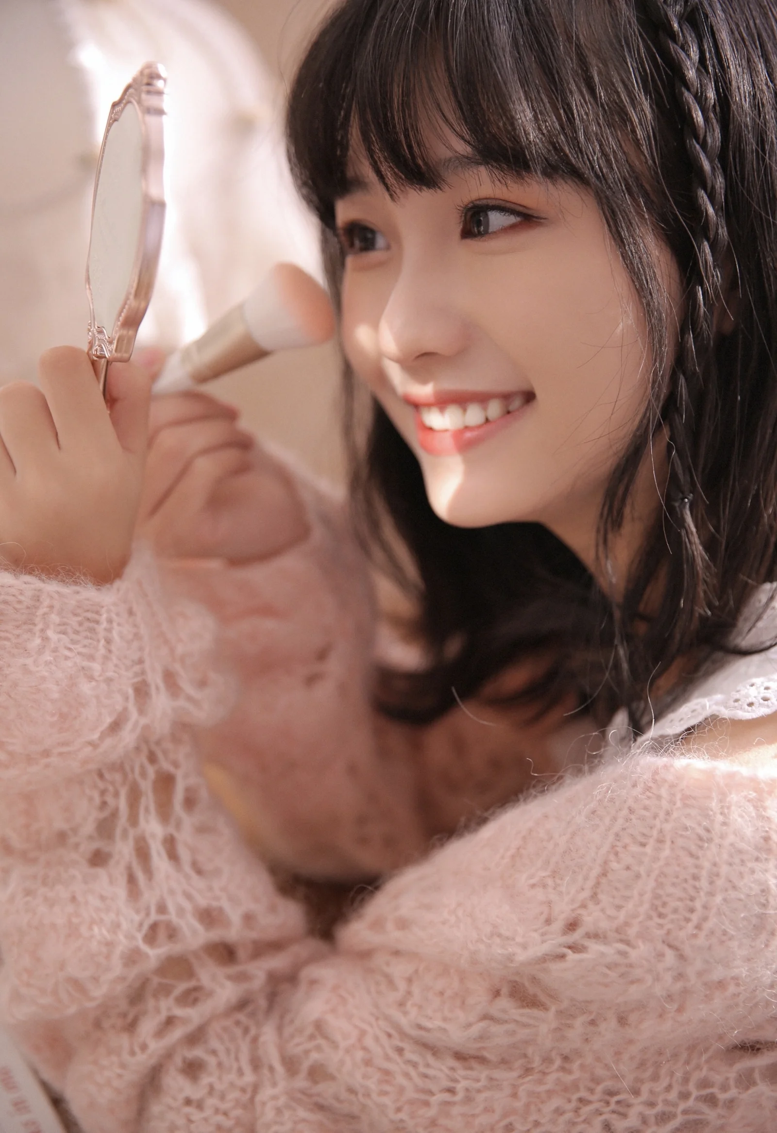 긴 머리의 여성이 거울과 메이크업 브러시를 들고 미소 지으며 화장을 고치고 있다.