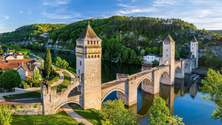 阳光下的石砌古桥横跨河流，桥塔、河畔村落与葱郁森林构成一幅开阔的风景画面。