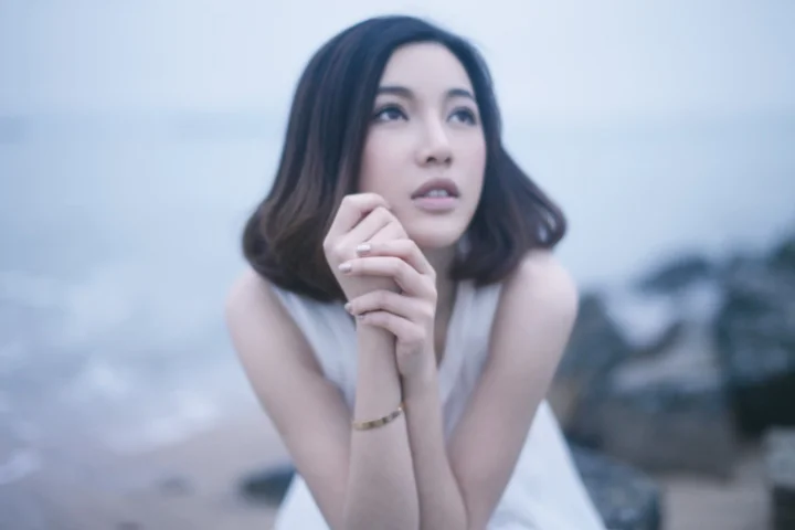 성인 여성이 두 손을 모은 채 부드러운 푸른 색조의 야외 배경 앞에서 먼 곳을 바라보고 있다.