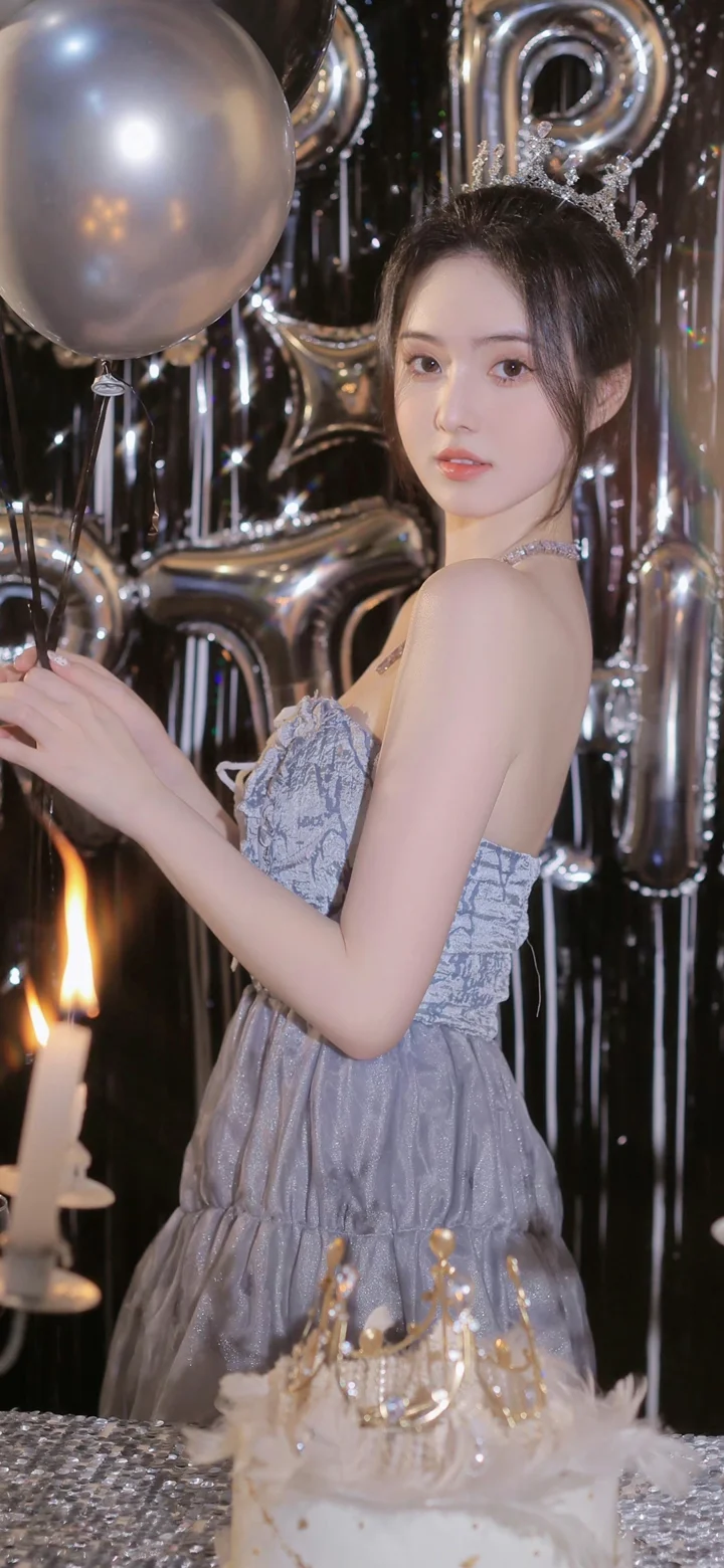 一位身穿银灰色礼服的女性站在银色气球和生日蛋糕前。