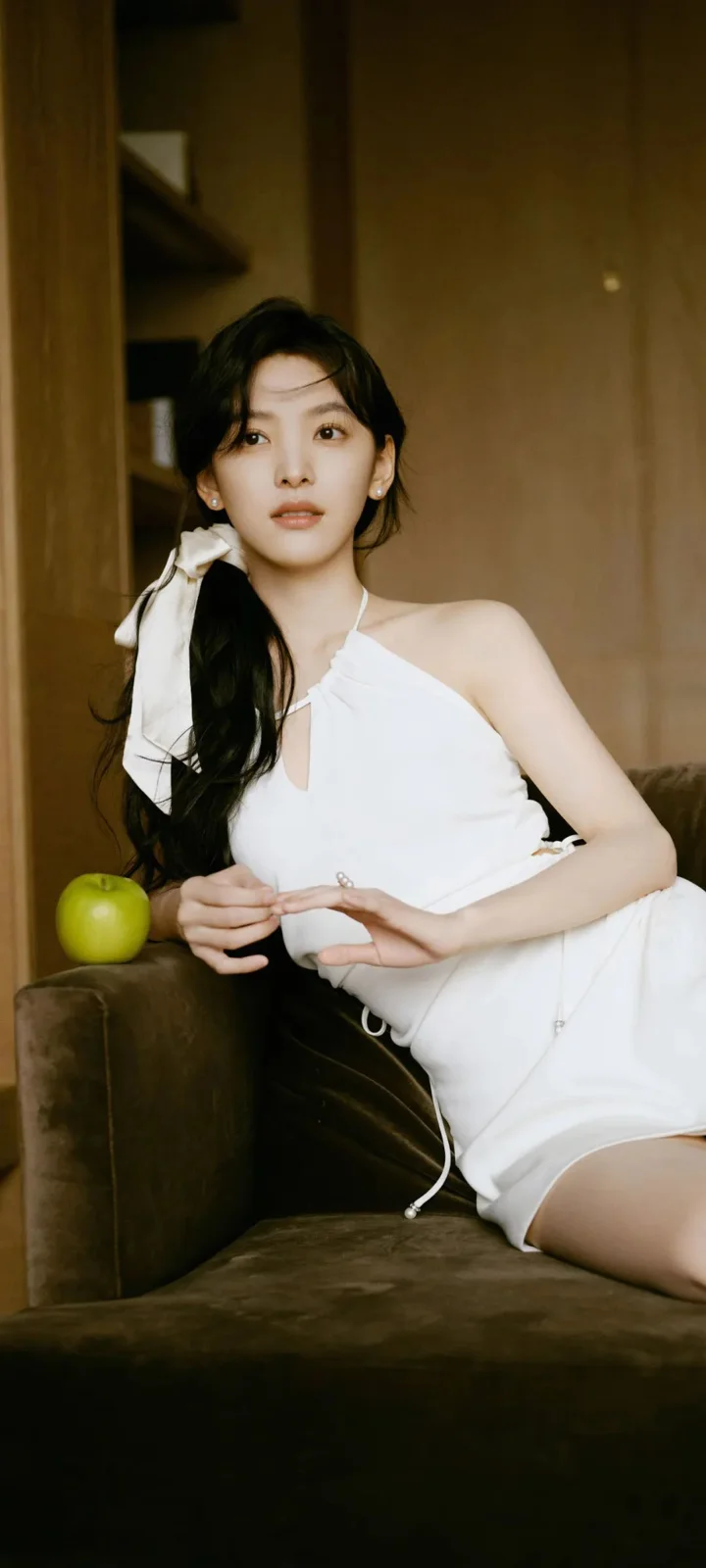 一名身穿白色连衣裙的成年女性坐在沙发上，旁边放着一只青苹果。
