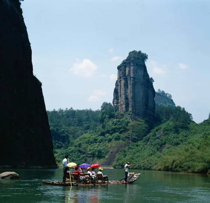 一座高耸的岩石山峰矗立在森林与河流之间，几个人乘坐竹筏漂行水面。