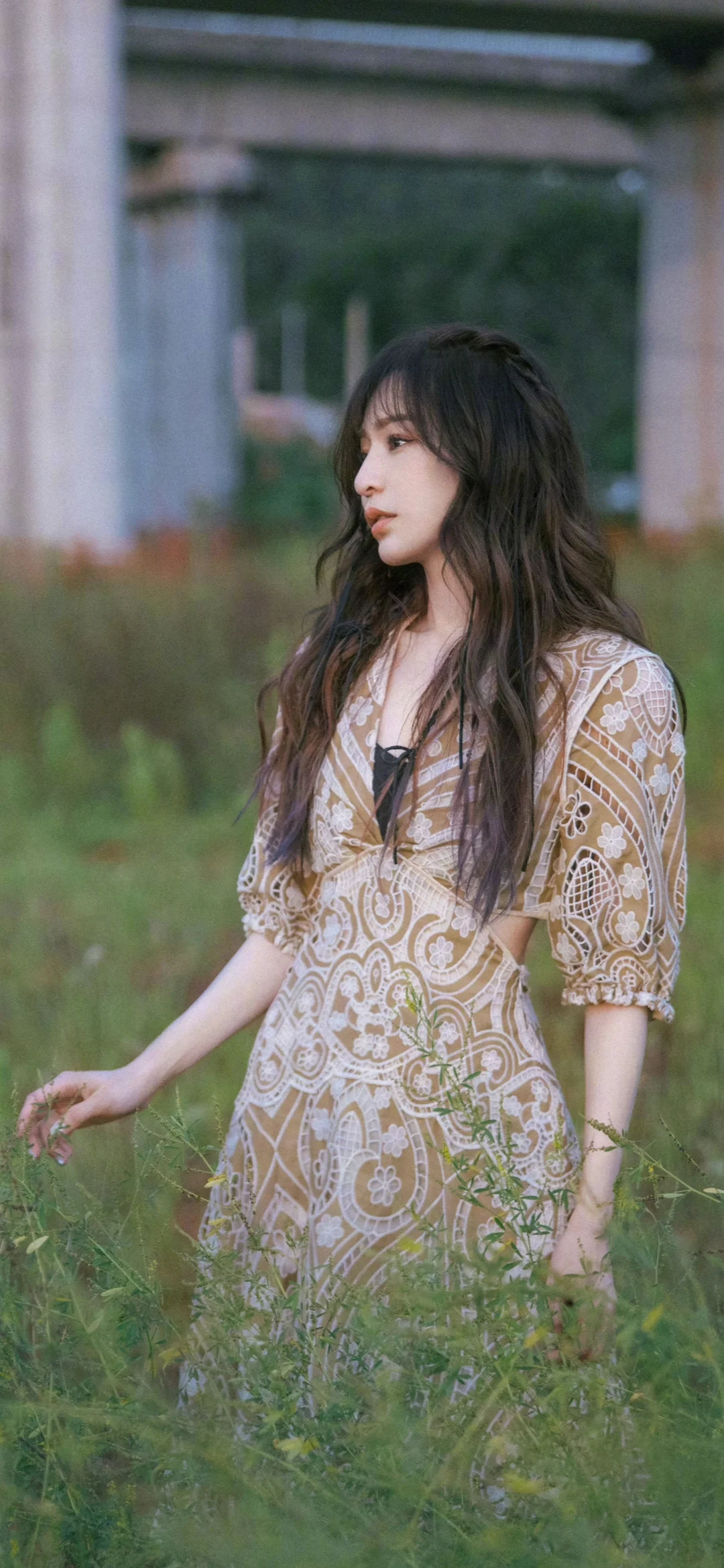 一名長髮女子穿著圖案連身裙站在草地上，身後是混凝土結構和樹木。