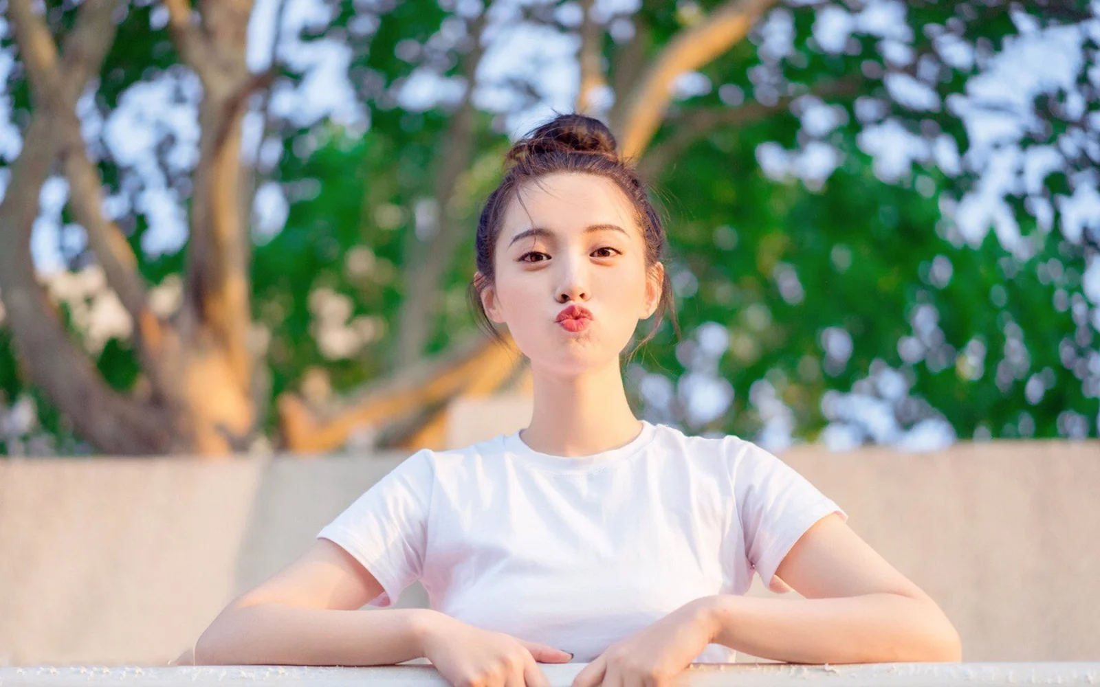 흰색 티셔츠를 입은 여성이 나무를 배경으로 카메라를 향해 입술을 오므리고 있습니다.