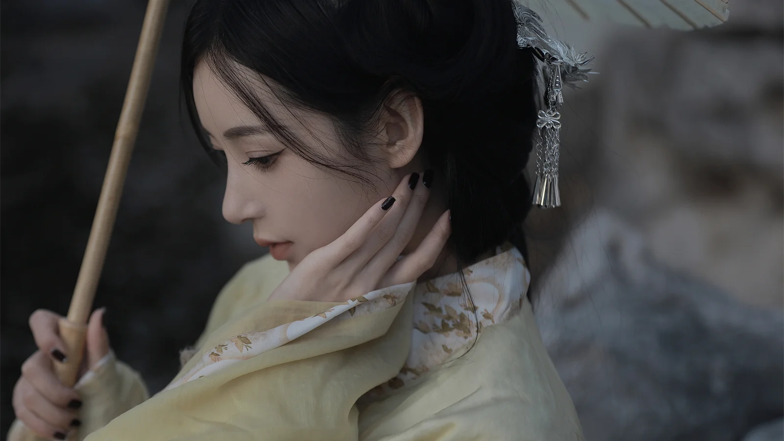한 여성이 옆모습으로 우산을 들고 밝은 색상의 무늬 옷을 입은 채 목 옆에 손가락을 대고 있다.