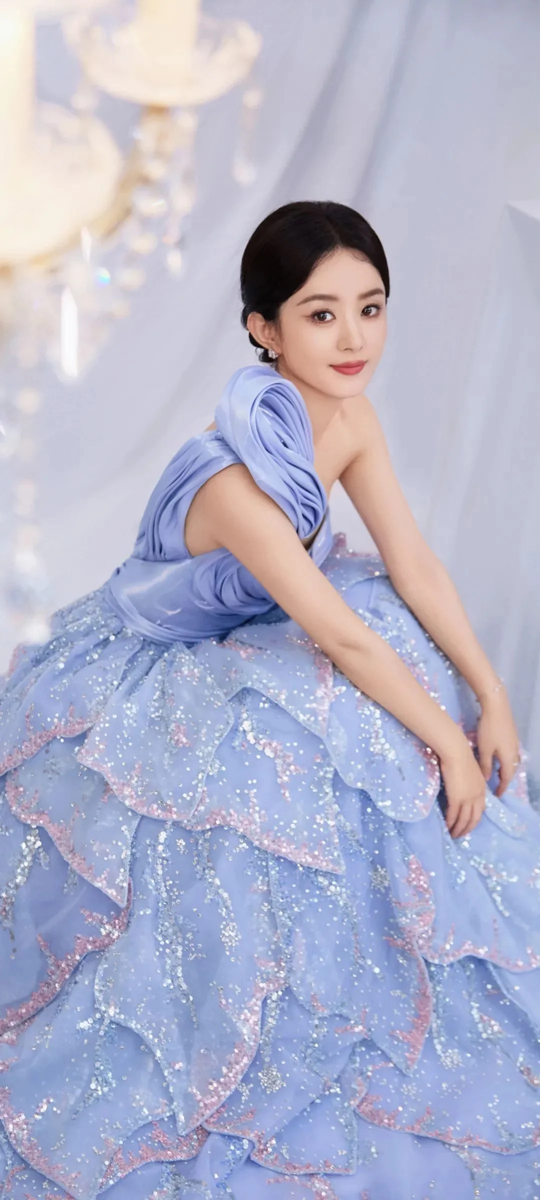 淡い青色の重ねドレスを着た成人女性が明るい背景の前に座っています。