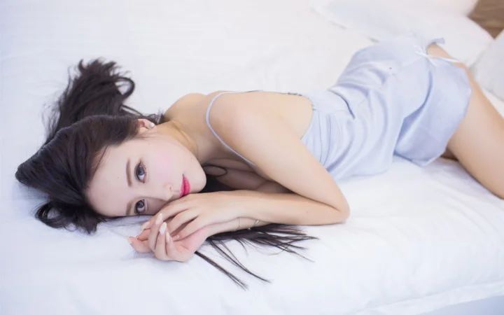 一名女性身穿浅色吊带裙侧躺在白色床上，面向镜头。