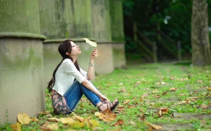 大人の女性が屋外の石柱のそばに座り、散った秋の葉の中で一枚の葉を手にしている。