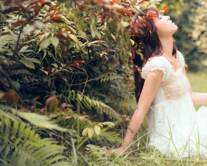 一名身穿白裙的女子站在茂密绿叶间仰头微笑。
