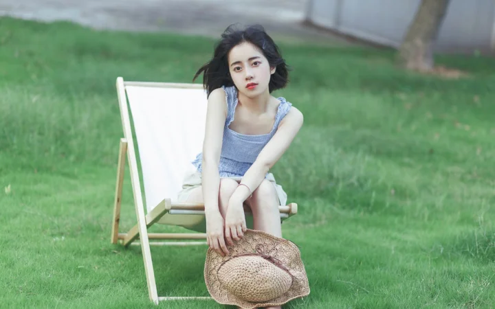 一名女性坐在草地上的折叠椅上，前景放着一顶草帽。